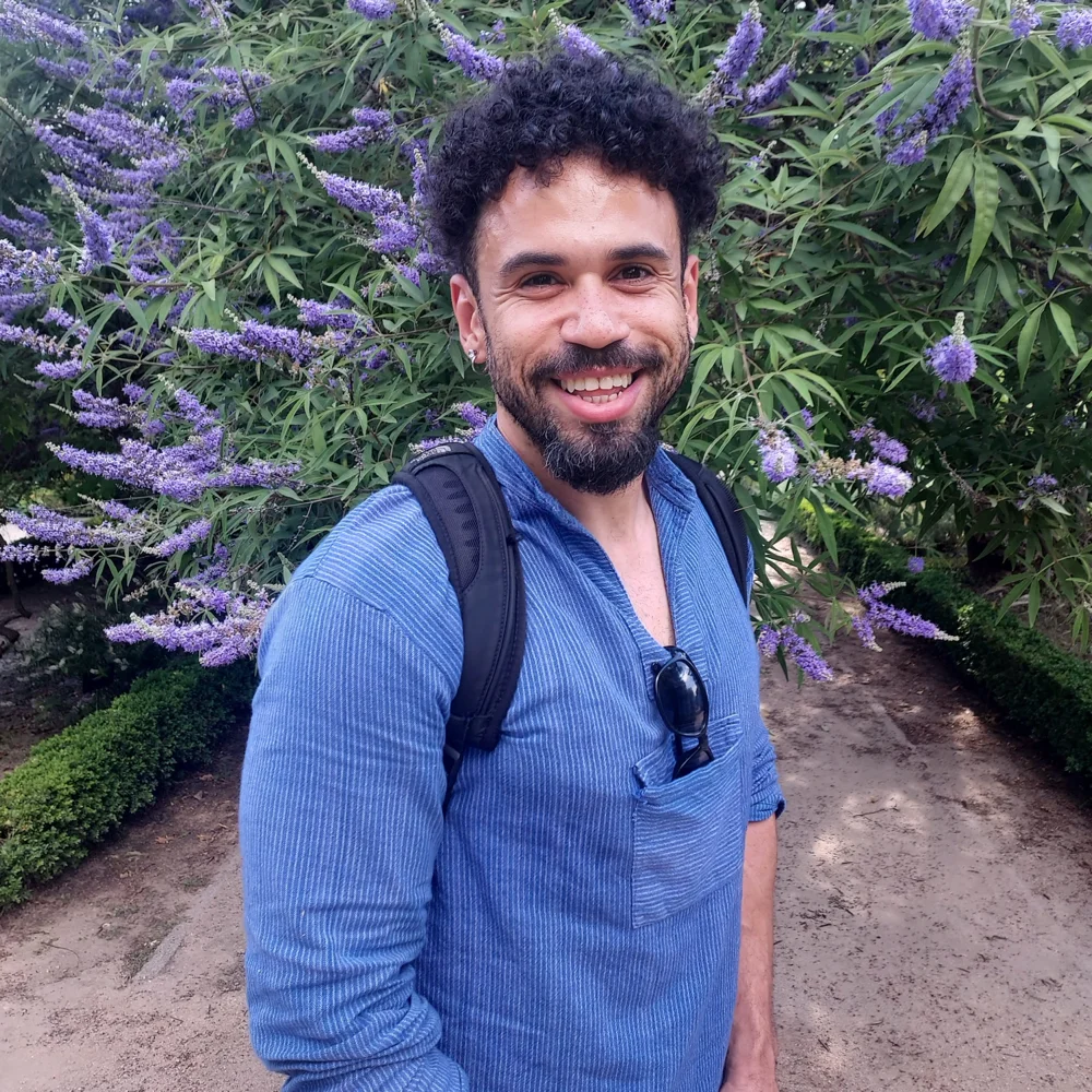 Luke Taylor smiling in front of a flowering lilac-purple tree, wearing a blue shirt and a backpack.